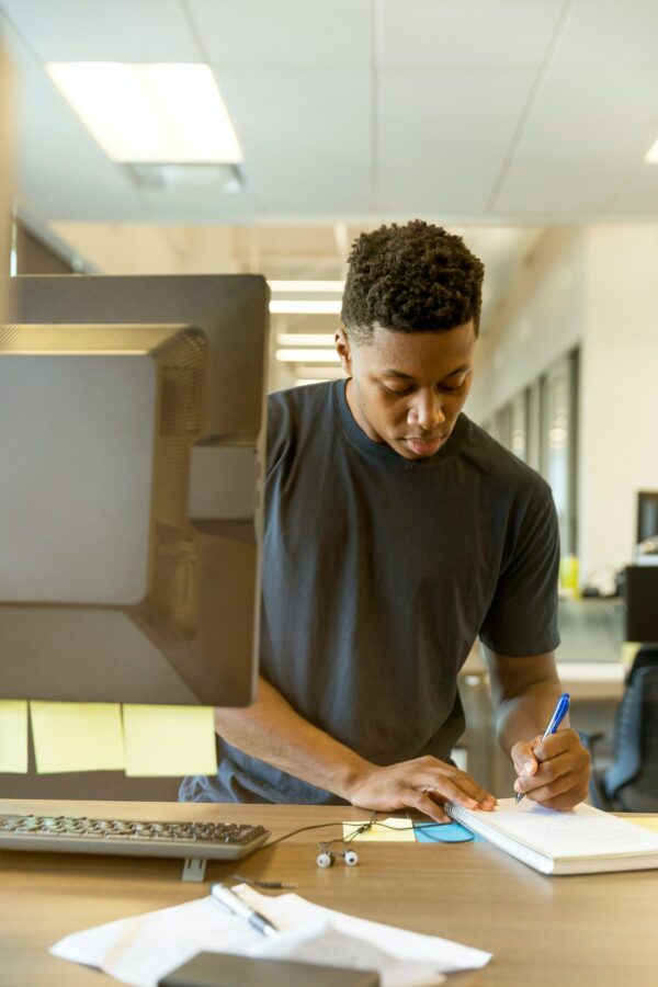 A young man stood at a desk making notes about key man insurance from a computer screen onto a notepage
